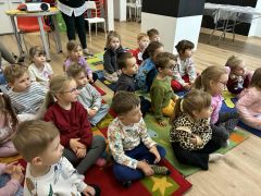 Dzieci w bibliotece siedzą na dywanie słuchają opowiadania. Pani czyta.